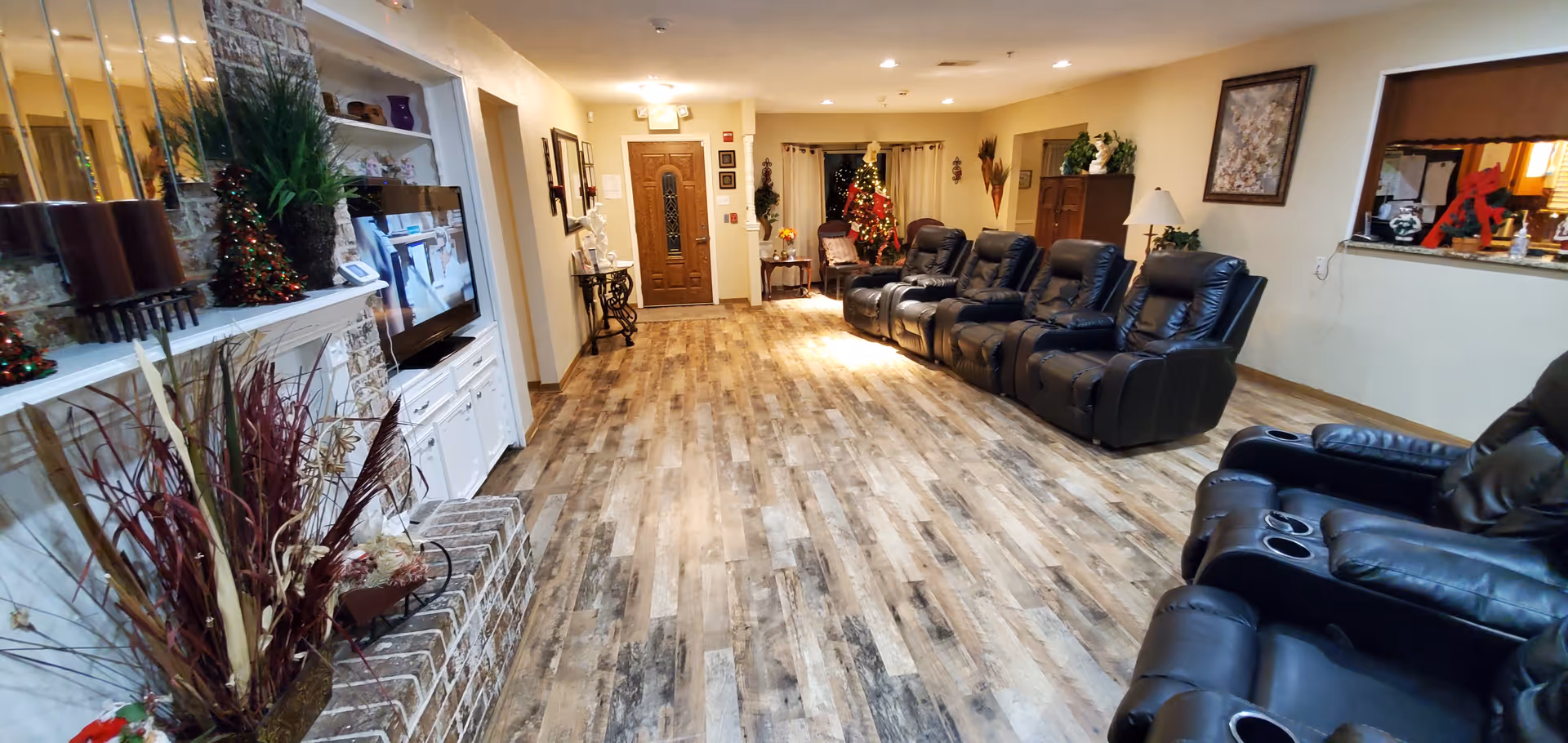A long, narrow living room area with several black leather recliner chairs lined up along the right side. On the left side, there is a brick fireplace with decorative plants and a TV mounted above a white cabinet. The room has wood-patterned flooring and light-colored walls with framed pictures and a Christmas tree decorated in the background near the entrance door.