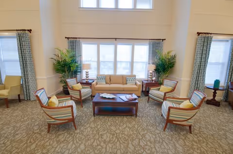 A bright and spacious living room with a beige sofa, four striped armchairs arranged around a wooden coffee table, two large potted plants, and windows with blue patterned curtains letting in natural light.