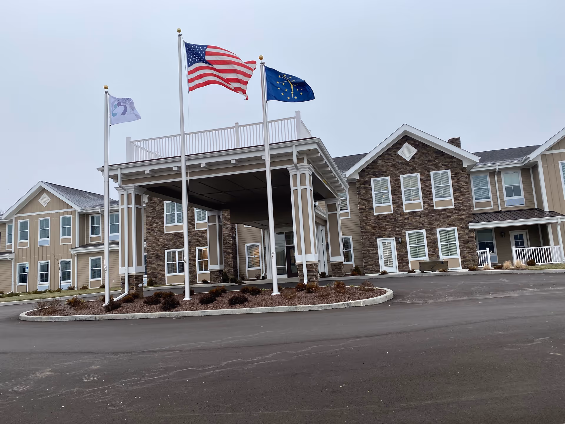 Front exterior of a two-story senior living building with a covered porte-cochère and three flagpoles flying flags.