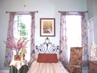 A cozy bedroom with a single bed featuring a decorative wrought iron headboard, a striped bedspread, and a red pillow. The room has two tall windows with floral curtains, a framed painting between the windows, a side table with a lamp, a framed photo, and a vase with pink flowers. There is also a floral upholstered chair and a dresser with a decorative statue.
