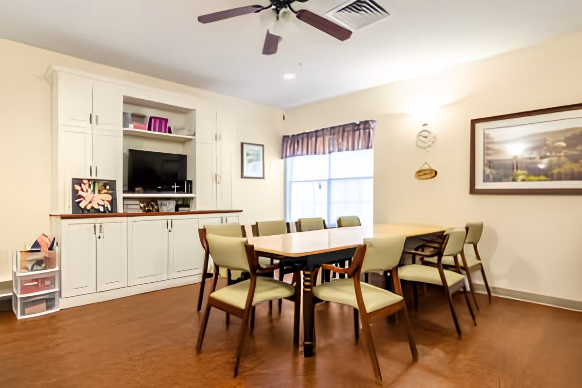 A community dining room with a long table surrounded by green-upholstered chairs, a built-in TV cabinet, and a window with valance.