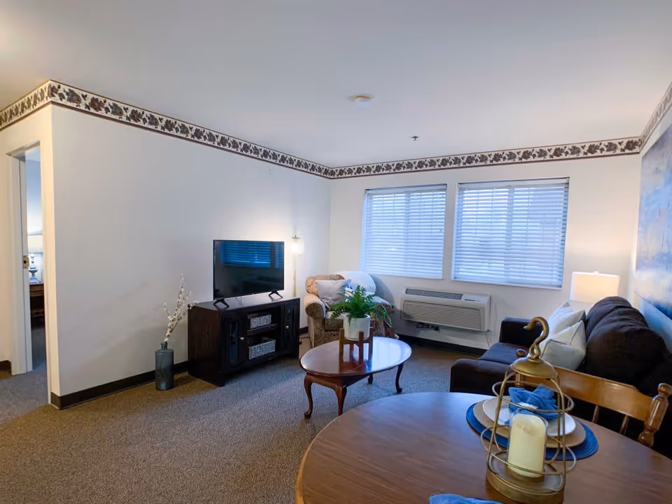 A cozy living room in an assisted living facility featuring a dark brown sofa with white pillows, a wooden coffee table with a potted plant, an armchair, a TV on a dark wooden stand, and a round dining table set with plates and a decorative candle holder. The room has beige walls with a floral border near the ceiling, two windows with blinds, and a wall-mounted air conditioning unit.