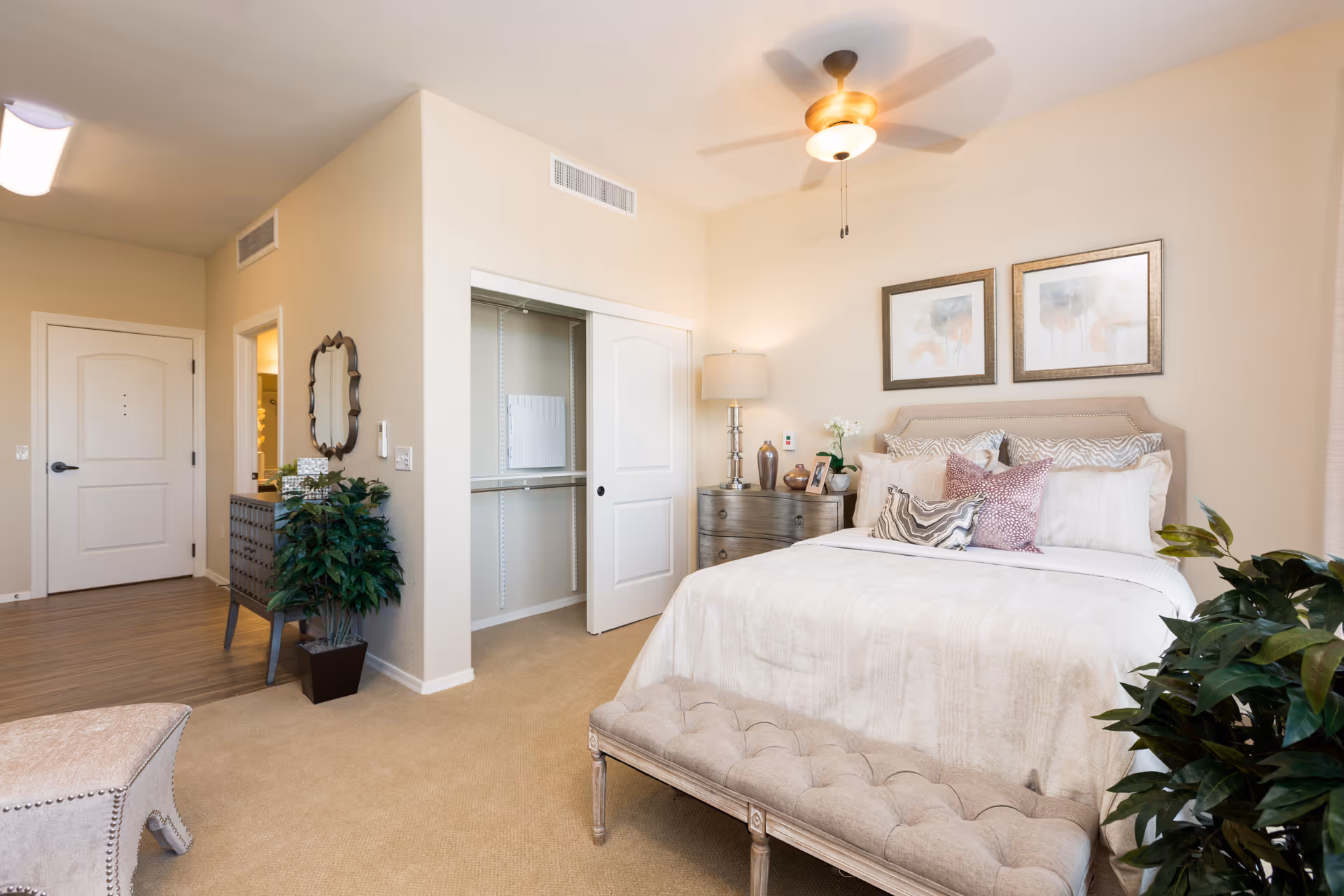 A bright and cozy bedroom in a senior living facility featuring a neatly made bed with multiple decorative pillows, a cushioned bench at the foot of the bed, a nightstand with a lamp and decorative items, two framed artworks on the wall above the bed, a ceiling fan with light, an open closet with sliding doors, a small dresser with a mirror, and potted plants adding greenery to the space.