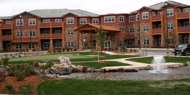 Exterior view of a multi-story senior living facility with stone and wood accents, featuring a landscaped lawn, small pond with a fountain, and paved walkways in front of the building.