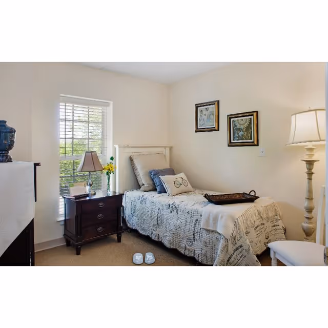 A small, cozy bedroom with a single bed covered in a patterned quilt and multiple pillows. Next to the bed is a dark wooden nightstand with a lamp, a vase with yellow flowers, and a small framed picture. There is a window with white blinds letting in natural light. On the wall above the bed are two framed artworks. A tall floor lamp stands near the bed, and a pair of slippers is placed on the carpeted floor at the foot of the bed.