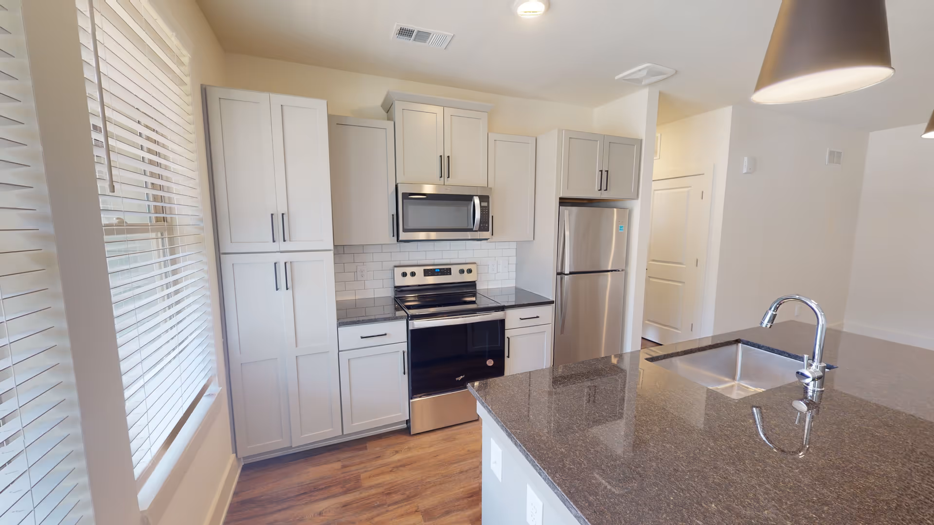 Modern kitchen with white cabinets, stainless steel appliances including a microwave, stove, and refrigerator, a large dark granite countertop island with a built-in sink and chrome faucet, and a window with white blinds letting in natural light.