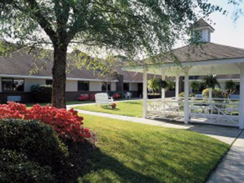 Courtyard with a white gazebo, benches, lawn, trees, and flowering shrubs in front of a single-story senior living building.