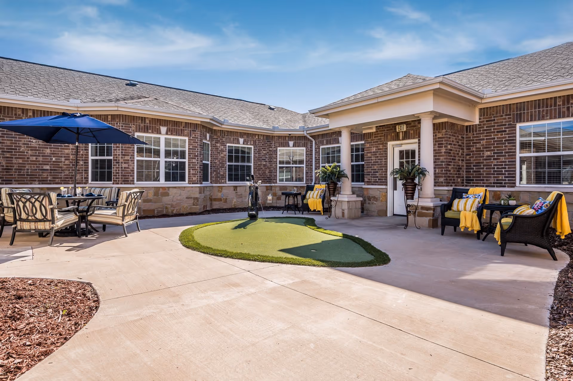 Outdoor patio area of a senior living facility with a small artificial putting green in the center. There are cushioned chairs with yellow blankets and pillows arranged around small tables, and a dining table with chairs under a blue umbrella. The building has brick walls and multiple windows under a clear blue sky.