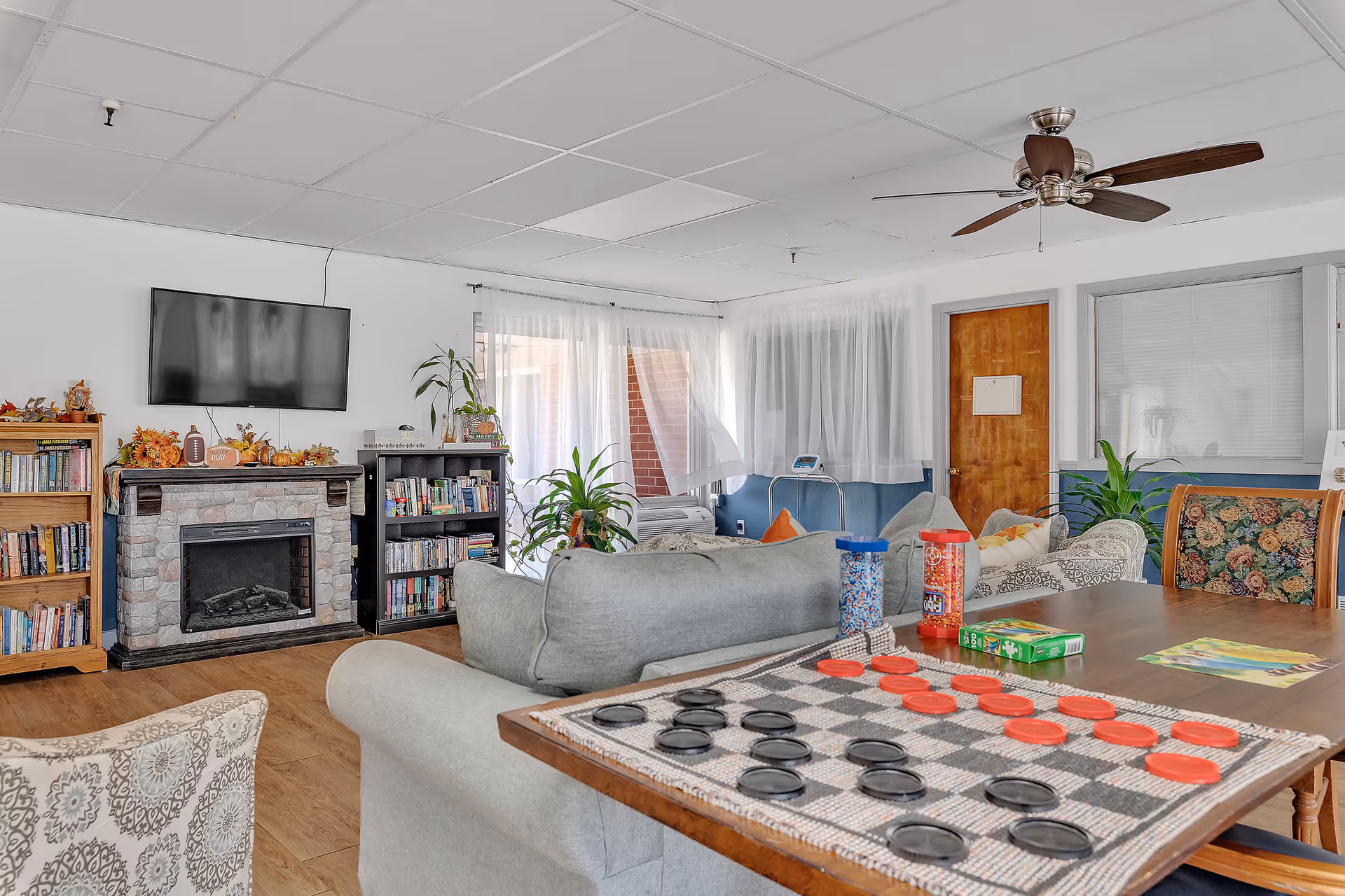 A bright communal living room with sofas, a wall-mounted TV above a faux fireplace, bookshelves, plants, and a checkers game laid out on a table.
