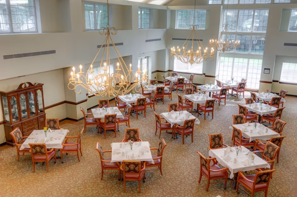 A spacious and well-lit dining room with multiple square tables covered with white tablecloths, each set with napkins, cups, and small flower vases. The room features large windows allowing natural light to fill the space, elegant chandeliers hanging from the ceiling, and upholstered chairs with floral patterns. A wooden cabinet with glass doors is visible against one wall.