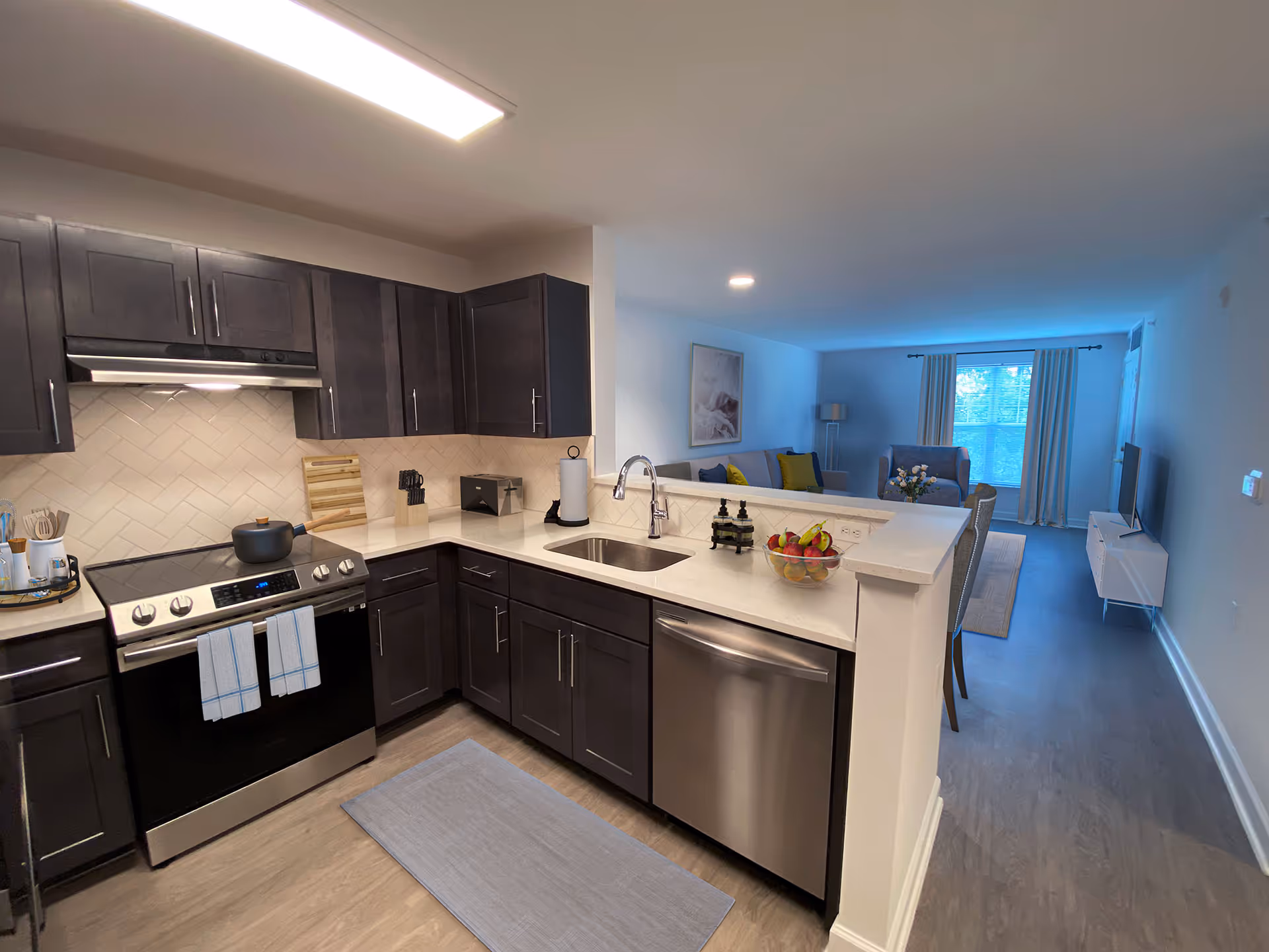 Open-plan kitchen with dark cabinets, stainless appliances, a white countertop island and a view into the living room.