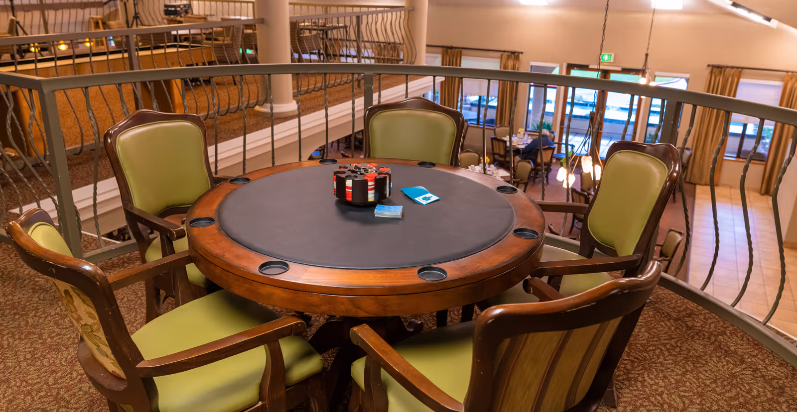 Round poker-style table with chips and cards surrounded by green upholstered chairs on an upper balcony overlooking a common area.