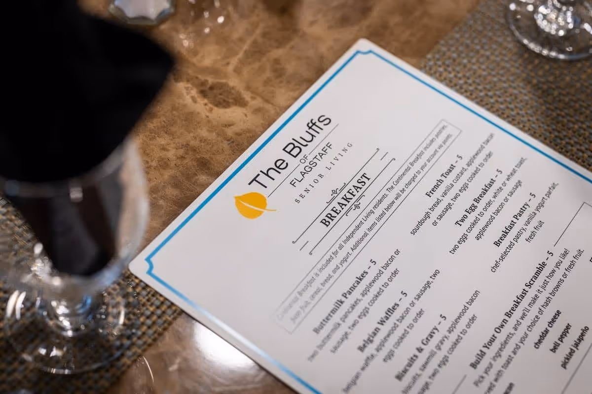 Close-up view of a breakfast menu from The Bluffs of Flagstaff Senior Living placed on a table with a woven placemat and a glass with a black napkin inside.