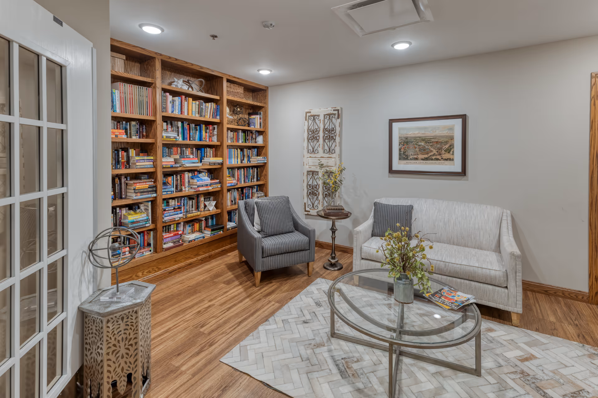 A cozy senior living common area with a wooden bookshelf filled with books, a gray armchair, a white loveseat with gray pillows, a round glass coffee table with a flower vase and magazines, a patterned rug, and framed artwork on the wall.