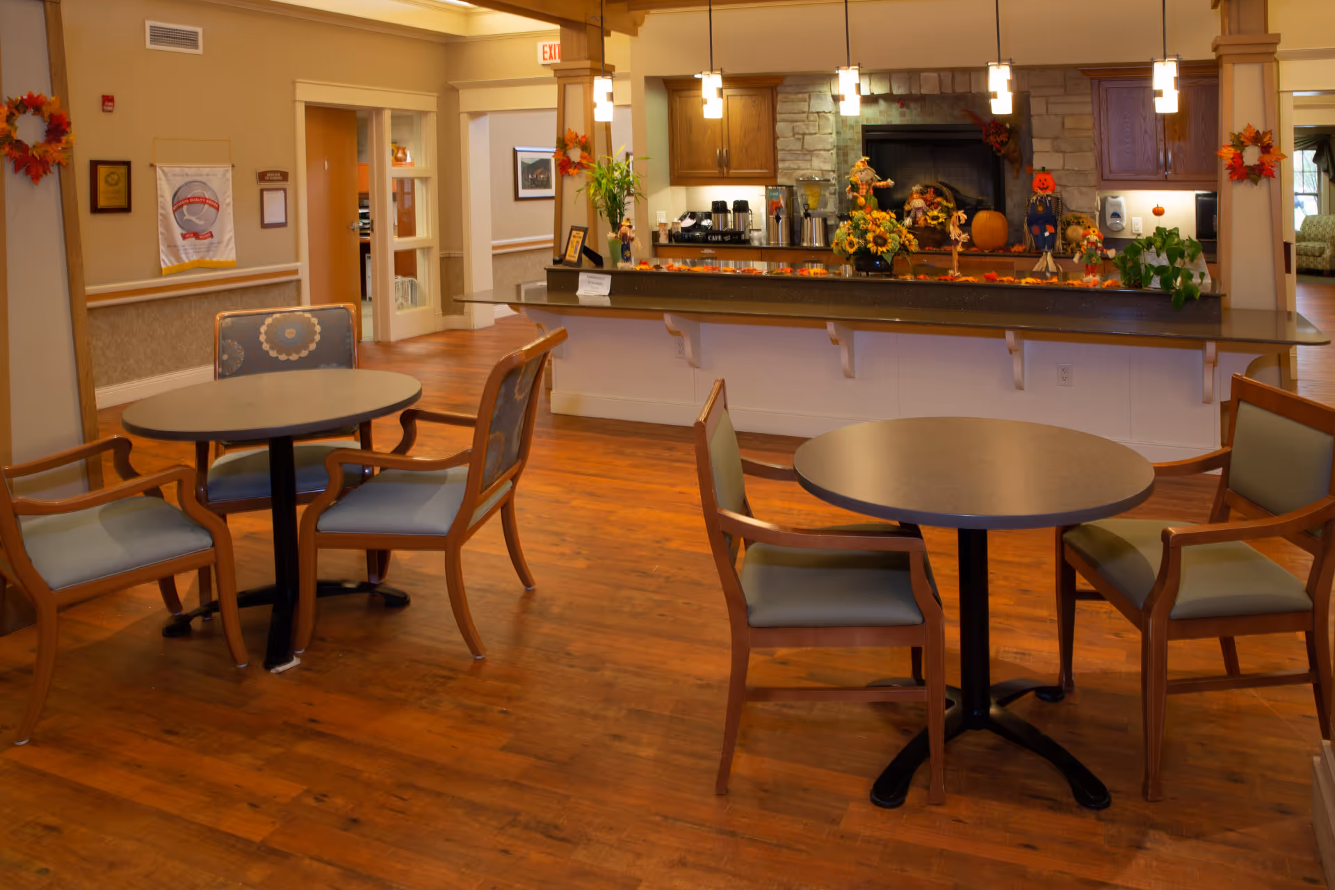 Interior view of a senior living facility dining area with round tables and wooden chairs. The room features hardwood floors, a counter with coffee and beverage dispensers, and autumn-themed decorations including pumpkins and fall wreaths. Warm lighting hangs from the ceiling, and there is a stone fireplace behind the counter.