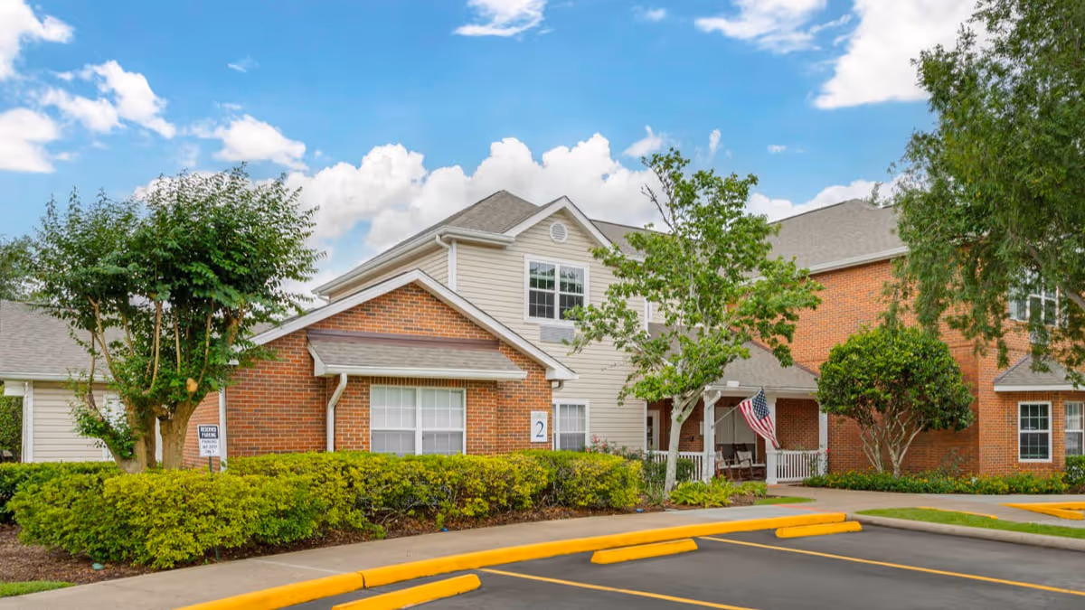 Front exterior of a brick-and-siding senior living building with a porch, American flag, landscaping, and parking curb.