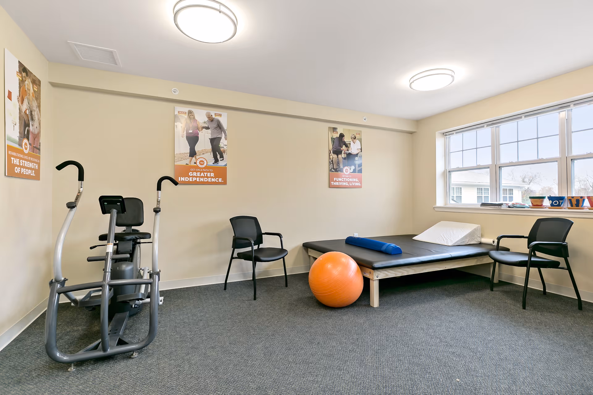 A small exercise room with a recumbent exercise bike, two black chairs, a padded exercise mat on a wooden frame with a wedge pillow and a blue bolster, and an orange exercise ball. The room has beige walls with three motivational posters, a large window letting in natural light, and a gray carpeted floor.