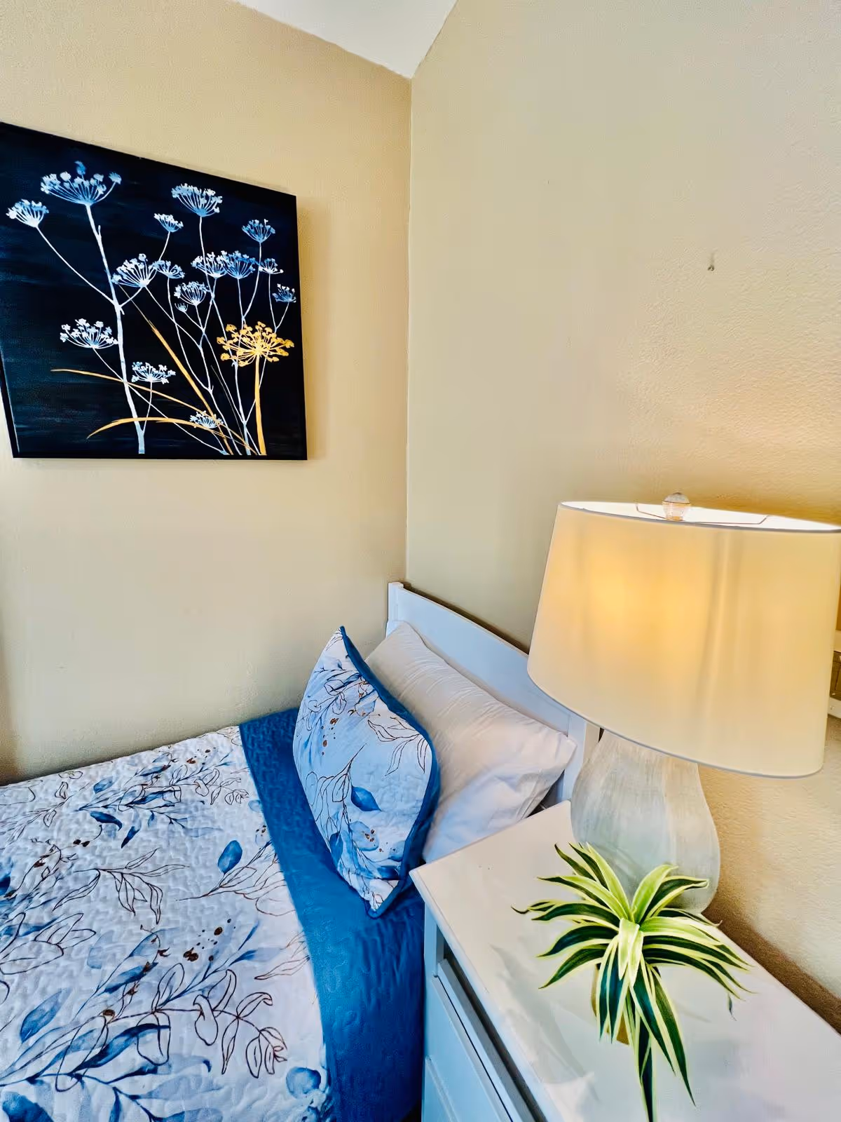 A cozy bedroom corner with a bed featuring blue and white floral bedding, a white pillow, a white nightstand with a green plant, and a lit table lamp. A framed artwork of white and yellow flowers on a dark background hangs on the beige wall above the bed.