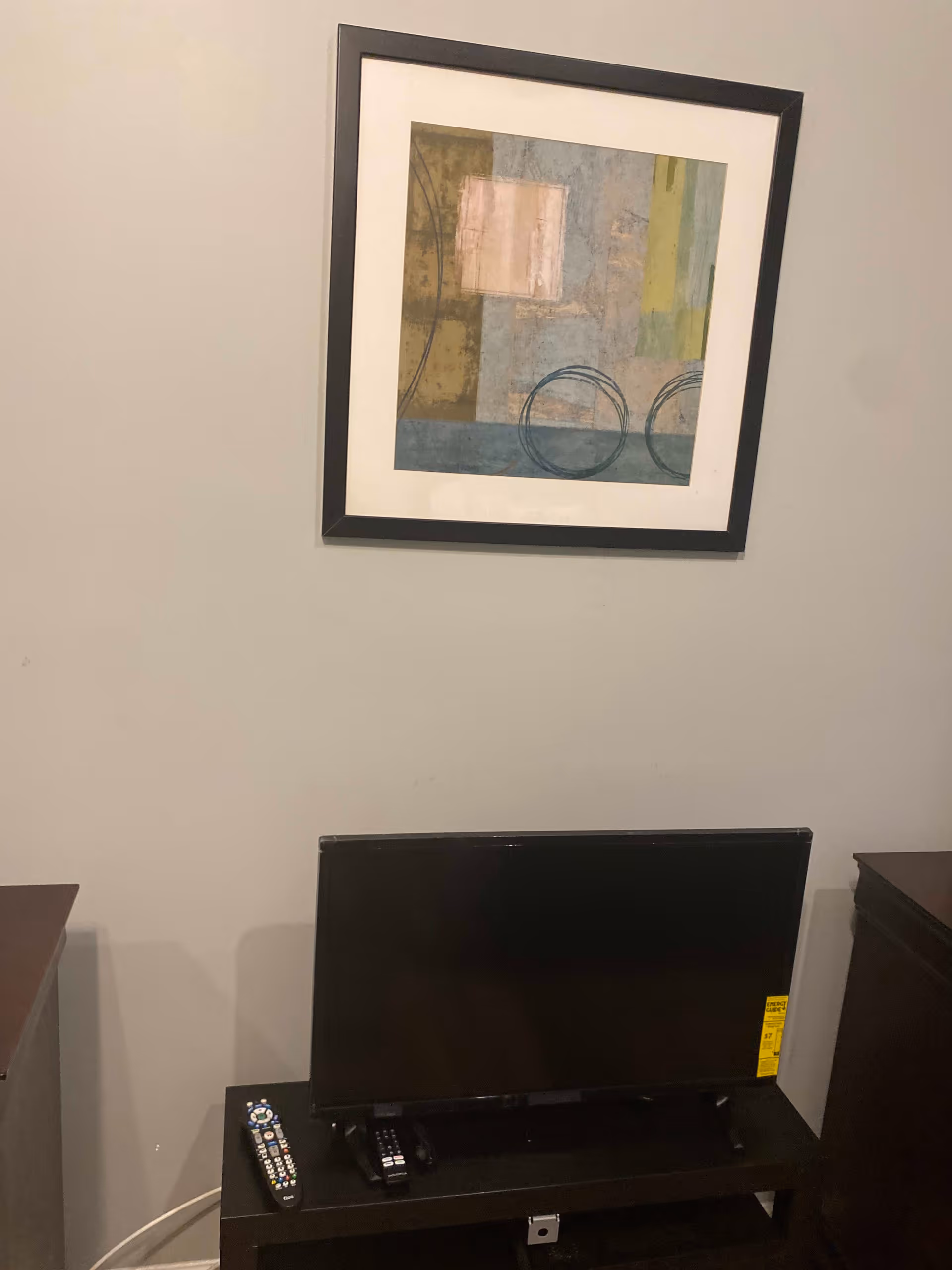 A small black flat-screen TV on a black stand with two remote controls placed on the stand. Above the TV, there is a framed abstract painting with geometric shapes in muted colors hanging on a plain light gray wall.