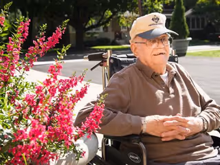 An elderly man wearing a beige cap and glasses sits in a wheelchair outdoors near vibrant pink flowers, with trees and a building in the background on a sunny day.