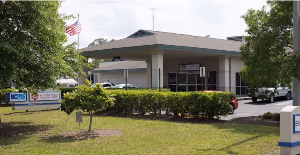 Exterior view of W Frank Wells Nursing Home building with a covered entrance, several parked cars, green bushes, trees, and an American flag on a flagpole.