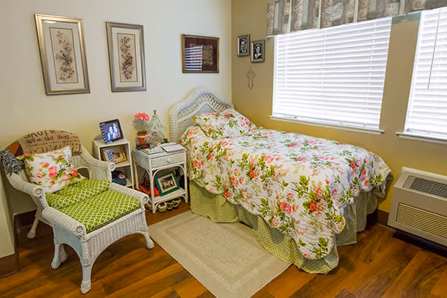 Small senior living bedroom with a single bed in floral bedding, a wicker chair, nightstand and windows with blinds.