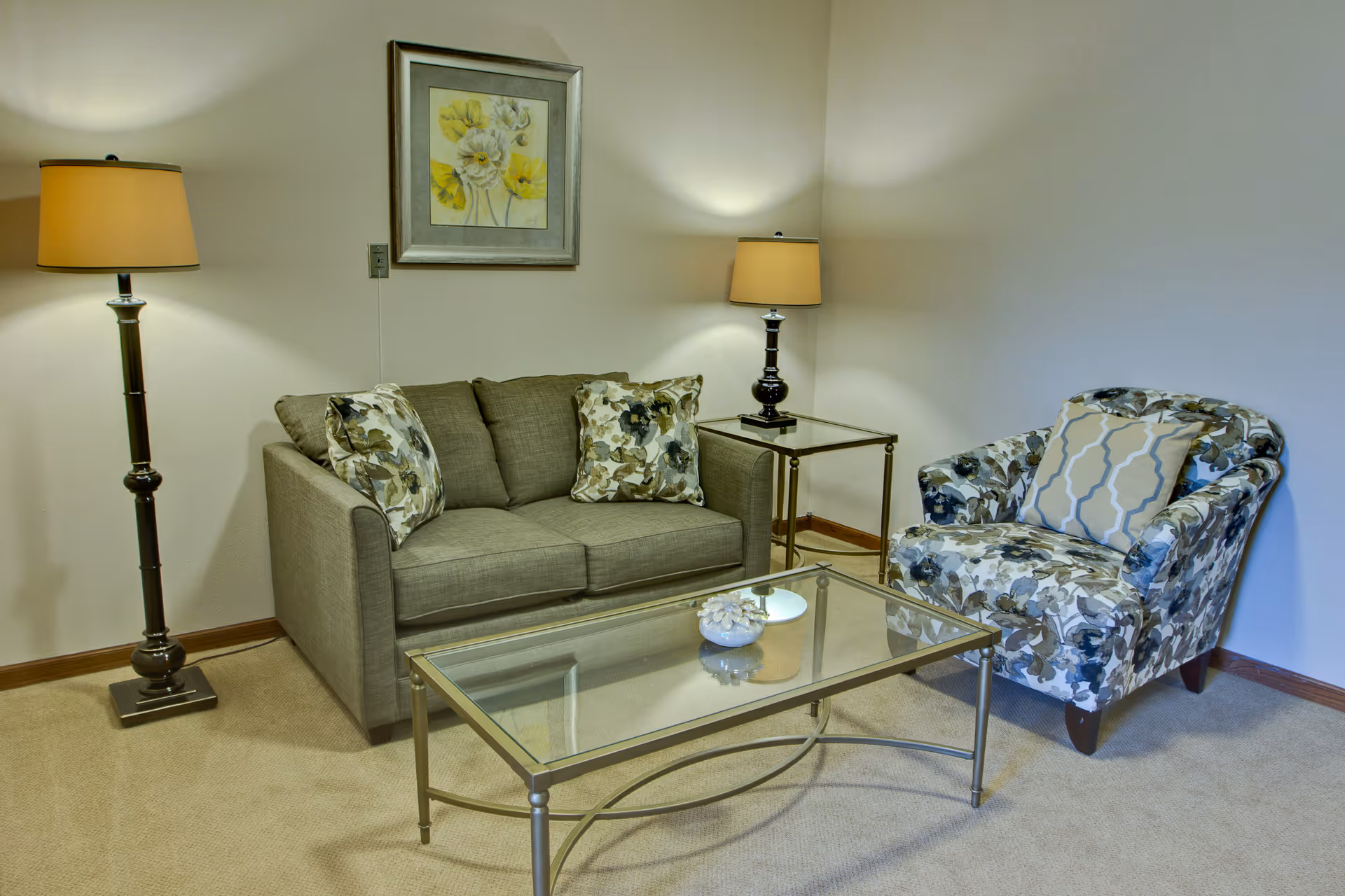 Small living room with a gray loveseat, patterned armchair, glass coffee table and two lamps.