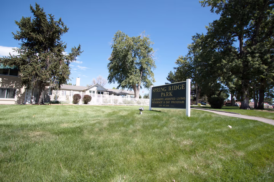 Outdoor view of Spring Ridge Park Assisted Living facility with a green lawn, trees, and a sign that reads 'Spring Ridge Park Alzheimer's Assisted Living Residence & Day Program'. The building is visible in the background under a clear blue sky.