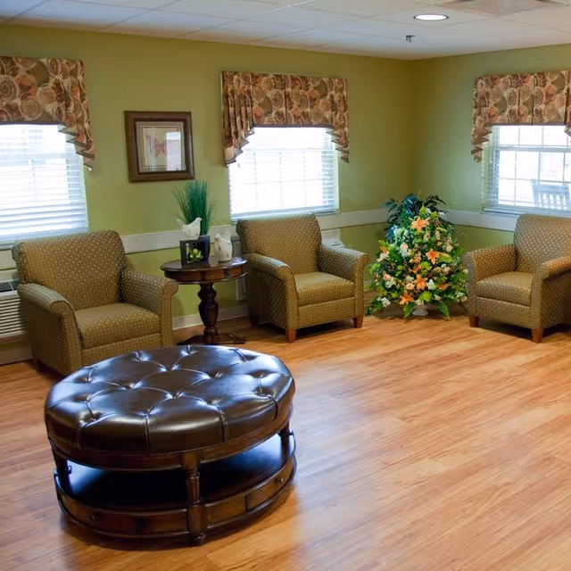 A cozy sitting area with three patterned armchairs arranged around a round, tufted leather ottoman on a wooden floor. The walls are painted light green and decorated with floral valances over three windows and a framed picture. A small round wooden table with decorative items and a floral arrangement is placed between two of the chairs.