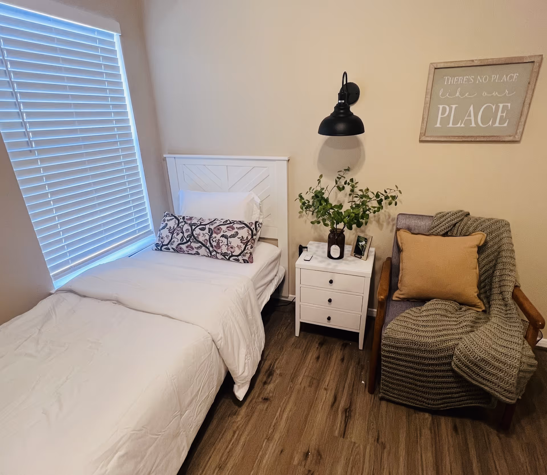 A cozy bedroom with a single bed dressed in white bedding and a decorative pillow. Next to the bed is a white nightstand with three drawers, holding a vase with green foliage, a small framed photo, and a candle. A gray armchair with a mustard yellow pillow and a knitted throw blanket is placed beside the nightstand. Above the chair, a wall-mounted black lamp and a framed sign that reads 'THERE'S NO PLACE like our PLACE' are visible. The room has wooden flooring and a window with white blinds.