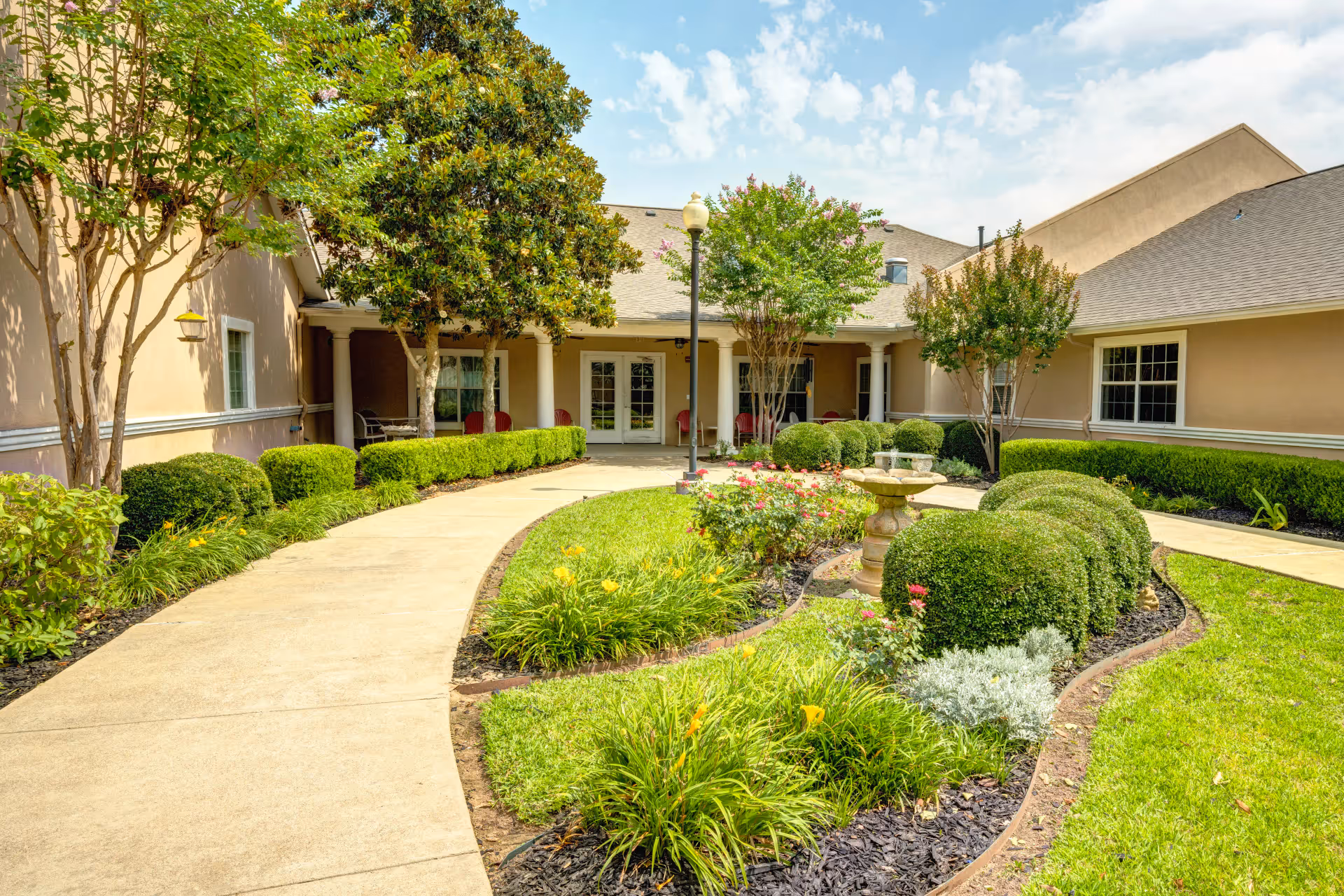 A landscaped courtyard at Dogwood Trails Assisted Living and Memory Care featuring a curved concrete walkway, manicured bushes, flowering plants, a stone birdbath, and several trees. The courtyard is surrounded by a beige building with white trim and columns supporting a covered patio area with red chairs.