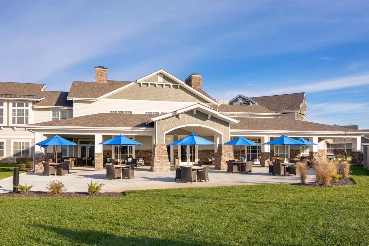 Outdoor patio area of American House Burlington Creek with multiple tables and chairs under blue umbrellas, adjacent to a large building with stone pillars and beige siding under a clear blue sky.