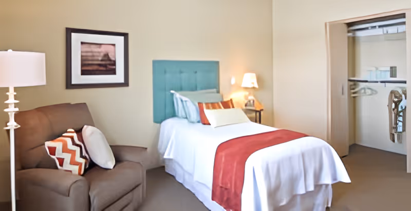 A neatly furnished single bedroom with a bed topped by a red runner and teal headboard, an armchair with patterned pillows, lamps, and an open closet.