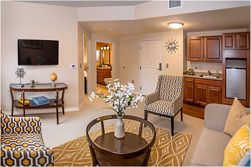 A cozy living room area in a senior living facility with a beige sofa, two patterned armchairs, a round glass coffee table with a vase of white flowers, a wall-mounted flat-screen TV, and a small kitchenette with wooden cabinets and a mini fridge. The room has a beige carpet and a decorative rug, with a view into a bathroom through an open door.