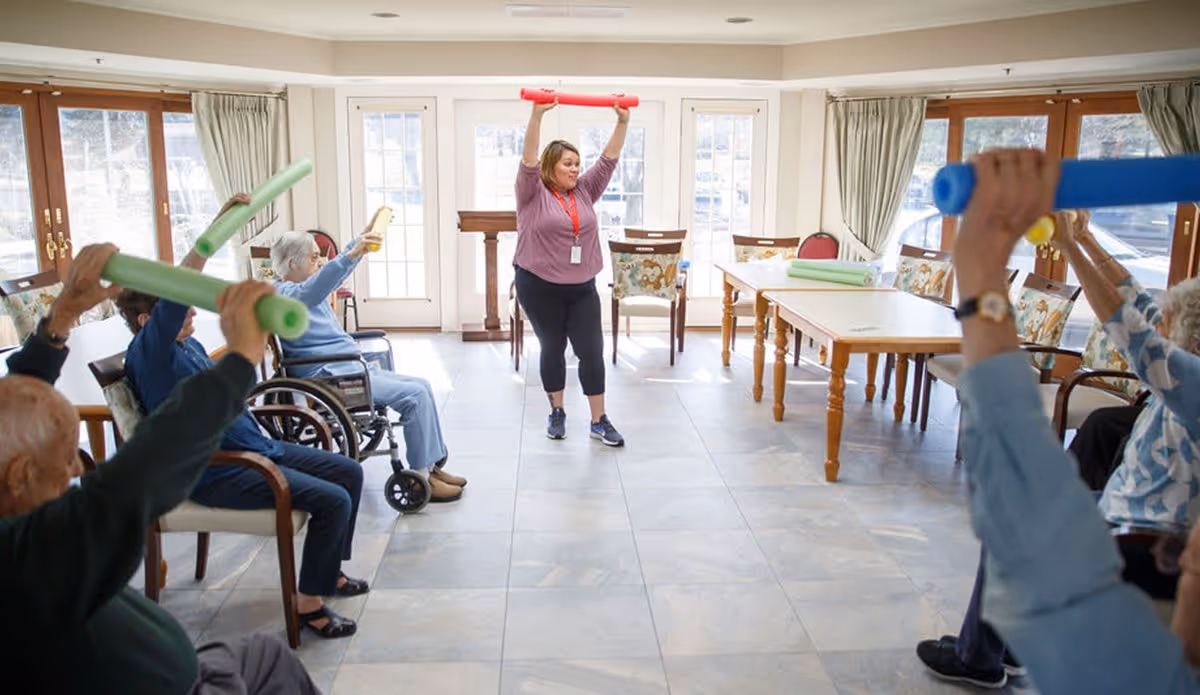 A group of elderly individuals seated in a spacious, well-lit room participate in a seated exercise class led by an instructor standing in front of them. The participants hold colorful foam noodles above their heads while following the instructor's movements. The room has large windows with curtains, wooden tables, and chairs arranged along the walls.