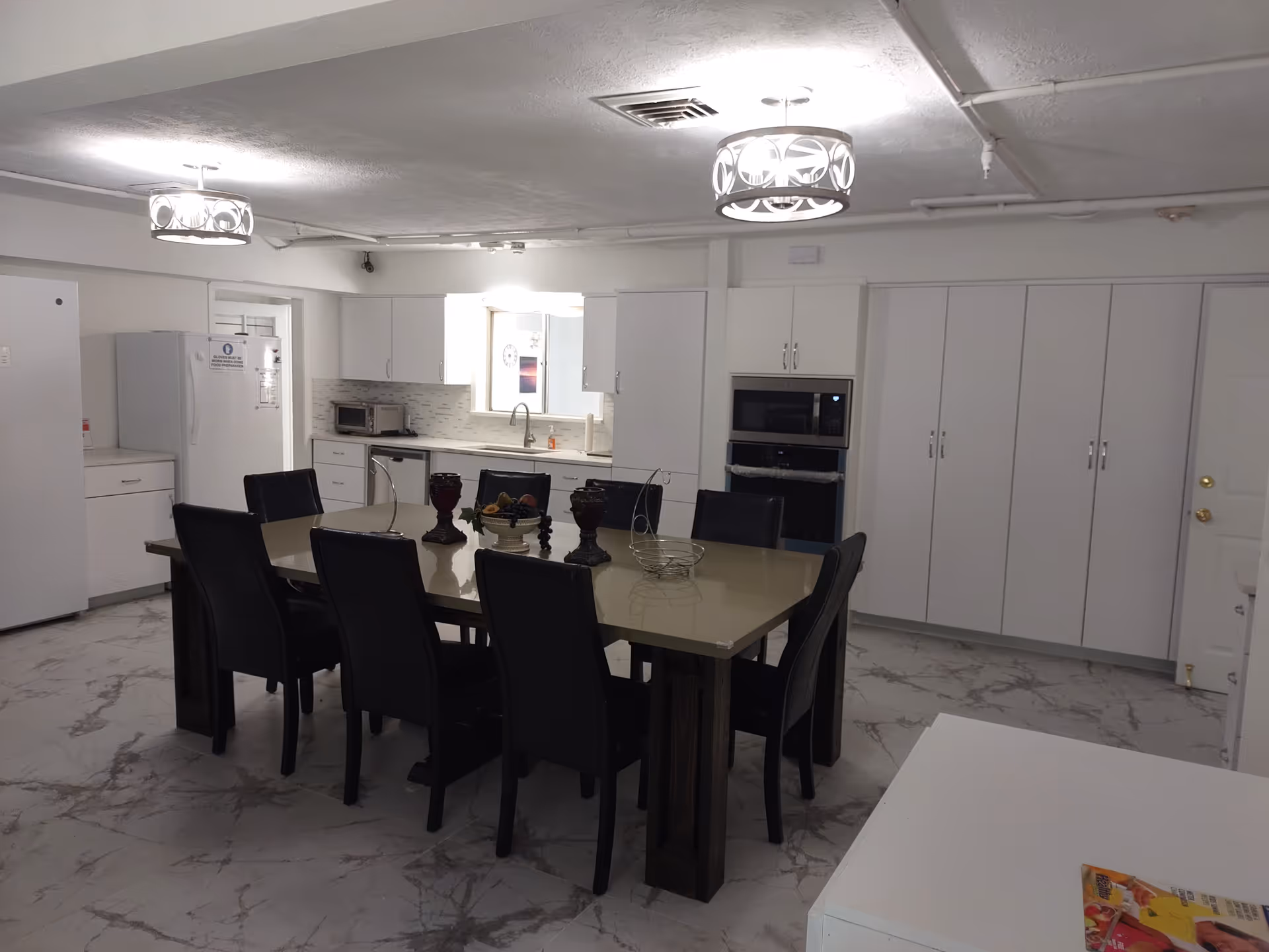 A spacious kitchen with a large dining table surrounded by eight black chairs. The kitchen features white cabinets, a refrigerator, microwave, oven, and a sink under a window. The floor has a marble-like pattern, and two modern ceiling lights illuminate the room.
