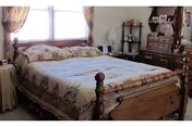 A cozy bedroom featuring a wooden bed with a patterned quilt, two windows with floral curtains behind the bed, a wooden dresser with a mirror, and a small shelving unit with decorative items.