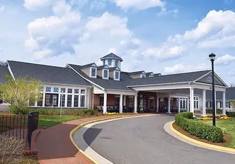 Exterior view of a single-story senior living facility building with a covered entrance, large windows, and a circular driveway. The sky is partly cloudy and there are landscaped bushes and a lamppost near the driveway.