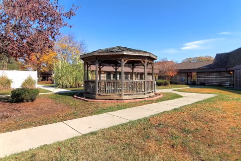 Outdoor area of Valley View Healthcare Center featuring a wooden gazebo surrounded by paved walkways, grass, and trees with autumn foliage under a clear blue sky.