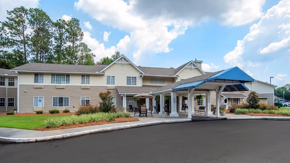 Exterior view of a two-story senior living facility with beige siding and white trim. The building has a covered entrance with a blue canopy and outdoor seating areas with tables and chairs. The surrounding area includes a well-maintained lawn, shrubs, and trees under a partly cloudy sky.