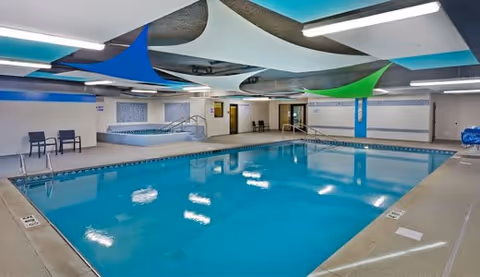 Indoor swimming pool area with clear blue water, surrounded by a tiled deck. There are a few chairs along the wall, a hot tub in the corner, and colorful triangular fabric shades hanging from the ceiling. The walls are decorated with blue and white tiles.