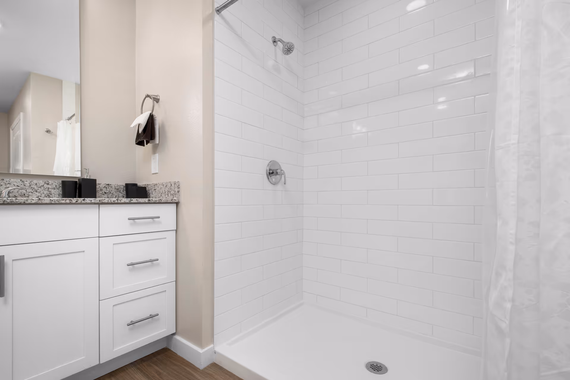 A clean bathroom featuring a white tiled shower with a silver showerhead and faucet. To the left, there is a white vanity with granite countertop, a large mirror, and a towel ring holding two towels. The floor is wood-style.