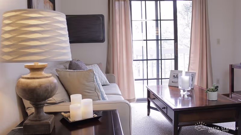 A cozy living room with a beige sofa adorned with multiple cushions, a wooden coffee table with candles and a small plant, a side table with a decorative lamp, and large sliding glass doors with pink curtains allowing natural light to fill the room.