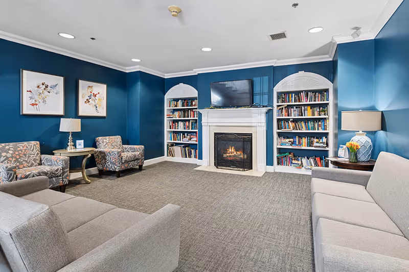 A cozy living room with blue walls featuring a white fireplace with a fire burning inside. Above the fireplace is a mounted flat-screen TV. On either side of the fireplace are built-in bookshelves filled with books. The room has two gray sofas facing each other and two patterned armchairs with a small round table and lamp between them. Another round table with a lamp and a vase of flowers is on the right side of the room. The floor is carpeted in a neutral tone.