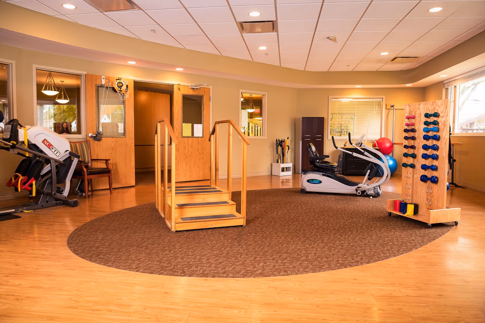 A rehabilitation or physical therapy room with wooden flooring and beige walls. The room contains exercise equipment including a recumbent bike, a small set of wooden stairs with handrails, a rack with various colored dumbbells, and exercise balls near the window. There are two wooden doors with glass panels and windows on the wall, allowing natural light to enter.