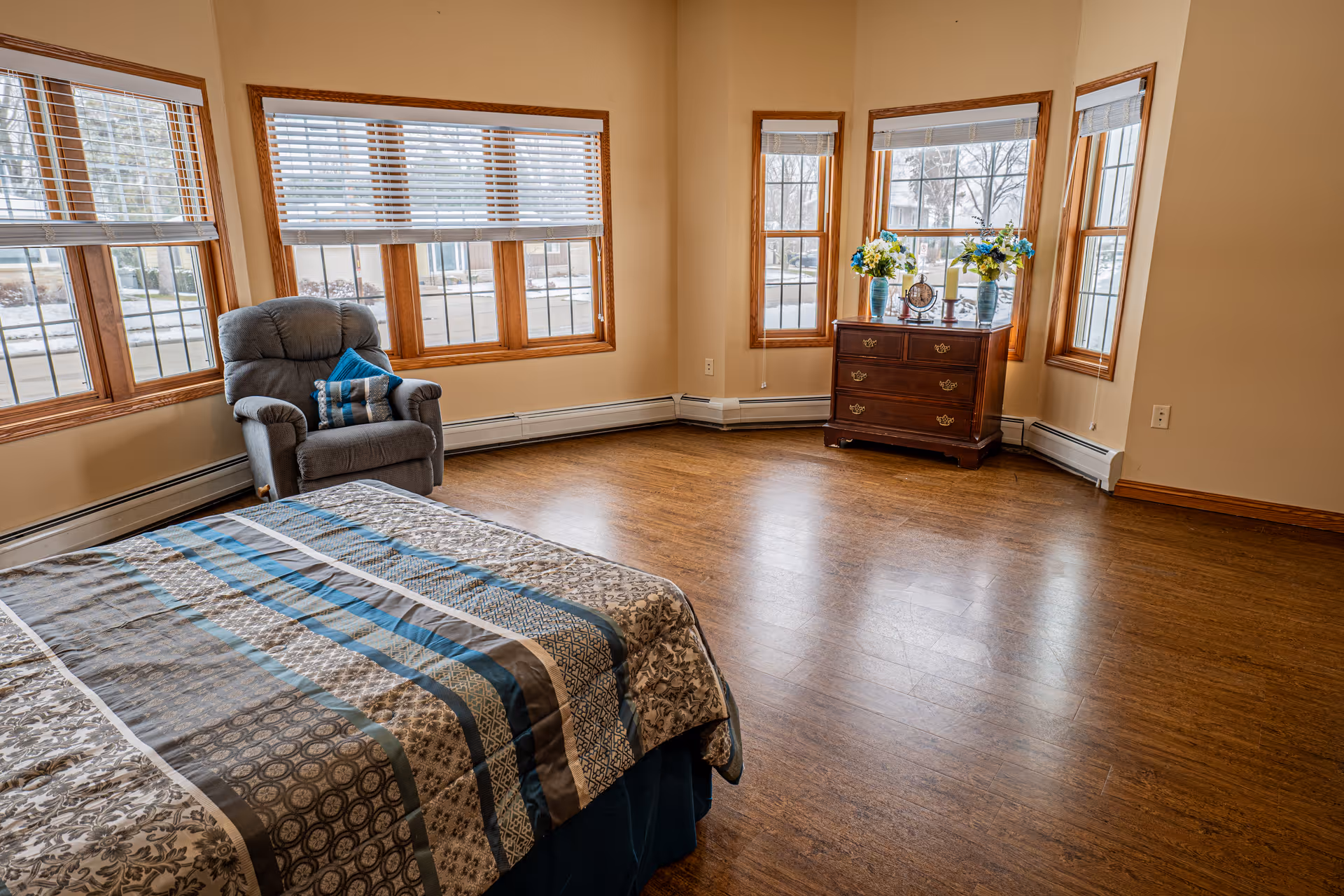 A bright bedroom with large windows featuring wooden frames and white blinds. The room has a bed with a patterned blue and gray bedspread, a gray recliner chair with a blue pillow, and a wooden dresser with decorative items including two vases with flowers, a clock, and a candle. The floor is wooden, and the walls are painted beige.