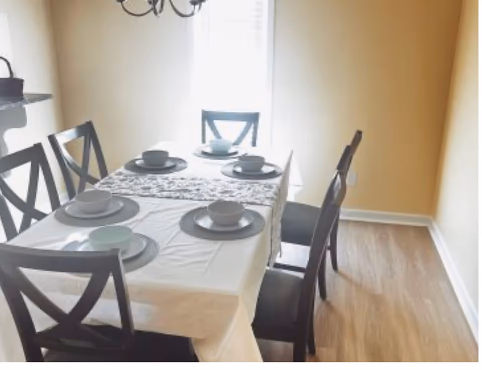 A dining room with a rectangular table covered with a white tablecloth and a decorative runner. The table is set with six place settings, each including a plate, bowl, and cup. Six dark wooden chairs with cross-back designs surround the table. The room has light-colored walls and wood flooring, with a window letting in natural light in the background.