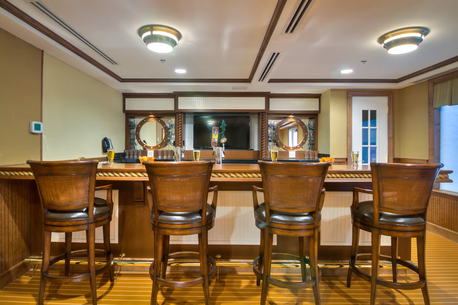 Wooden bar counter with four barstools in a well-lit common room featuring mirrors and a TV behind the bar.