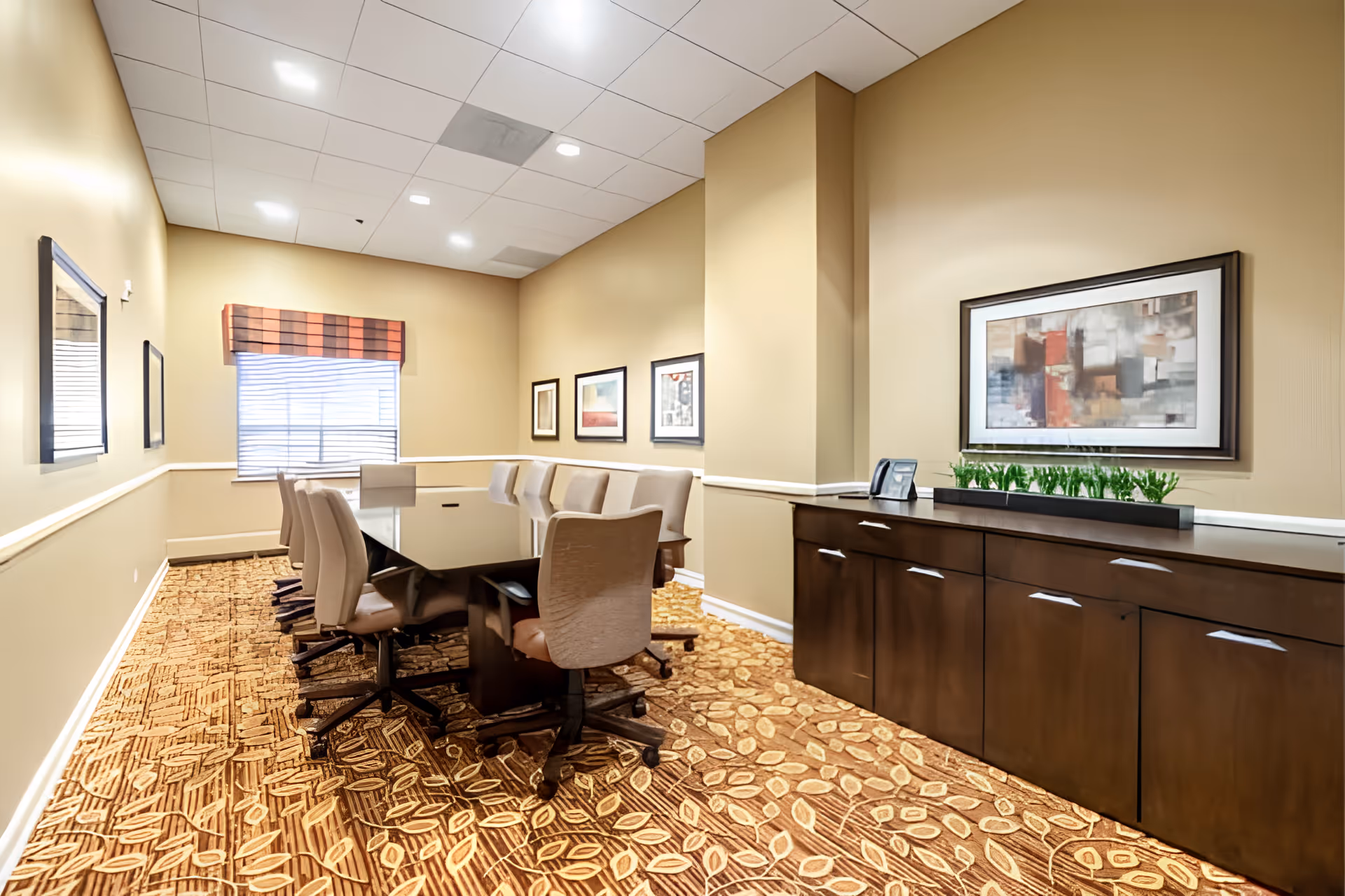 A well-lit conference room with a long rectangular table surrounded by swivel chairs, framed artwork on the walls, and a sideboard with decorative plants.