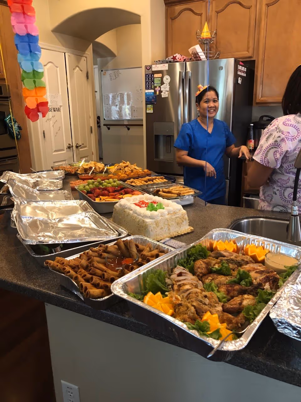 A kitchen counter filled with trays of food including grilled chicken, spring rolls, fresh fruit, and a decorated cake. Two women in scrubs are standing near a stainless steel refrigerator, one smiling and wearing a small party hat. The kitchen has wooden cabinets and a double sink.